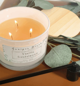 Candle with three lit wicks on the left says vanilla sandalwood on a white label. A leaf sits to the right of the candle on a wood table. A wooden candle lid sits in the upper right of the photo.